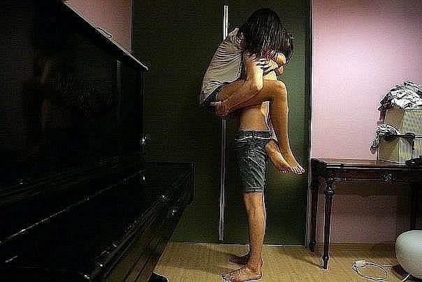 One person holds another in their arms in a room with a piano.